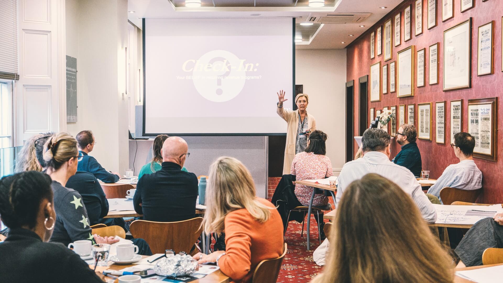 Jill S. Robinson stands presenting to an engaged audience in a meeting room decorated with framed artwork on deep red walls. She gestures while speaking, wearing a beige blazer. The room is filled with attendees seated at tables with coffee cups and materials, and a projection screen behind her displays the 'Check-In' title slide.