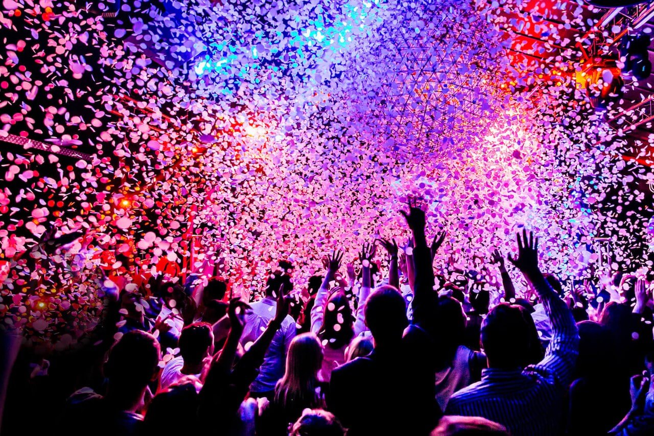 An excited crowd raise their hands as colorful confetti falls from above